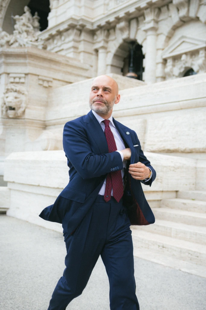 Mężczyzna w granatowym garniturze biznesowym, zapinający marynarkę, w białej koszuli i bordowym malinowym krawacie przed Sądem Najwyższym w Rzymie.