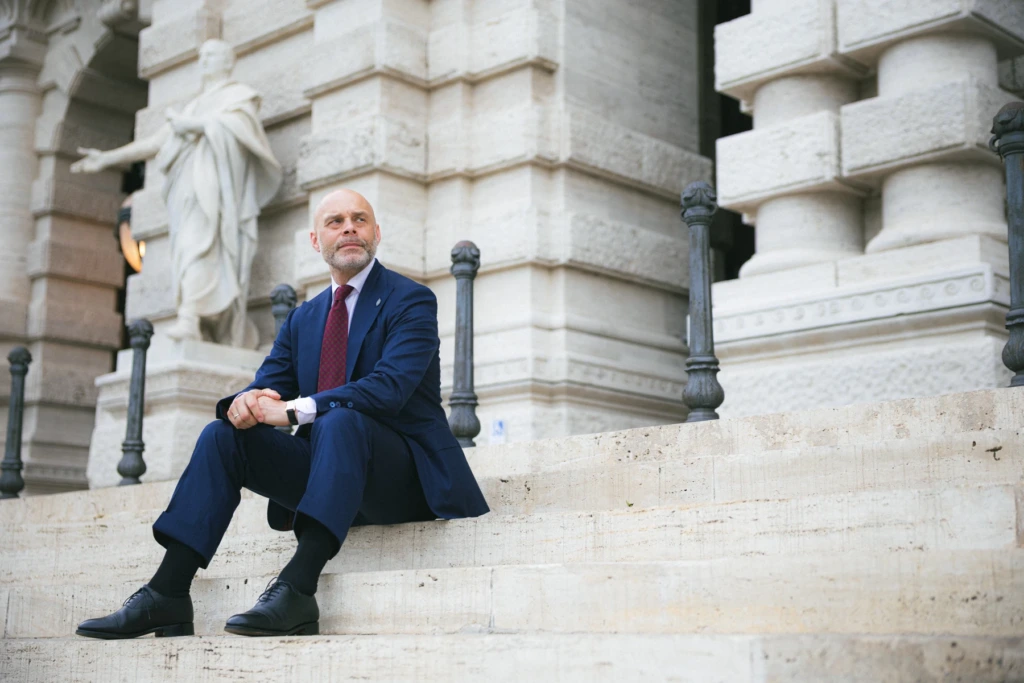 Mężczyzna w granatowym garniturze z wełny wysokoskrętnej siedzący na schodach przed Sądem Najwyższym w Rzymie.