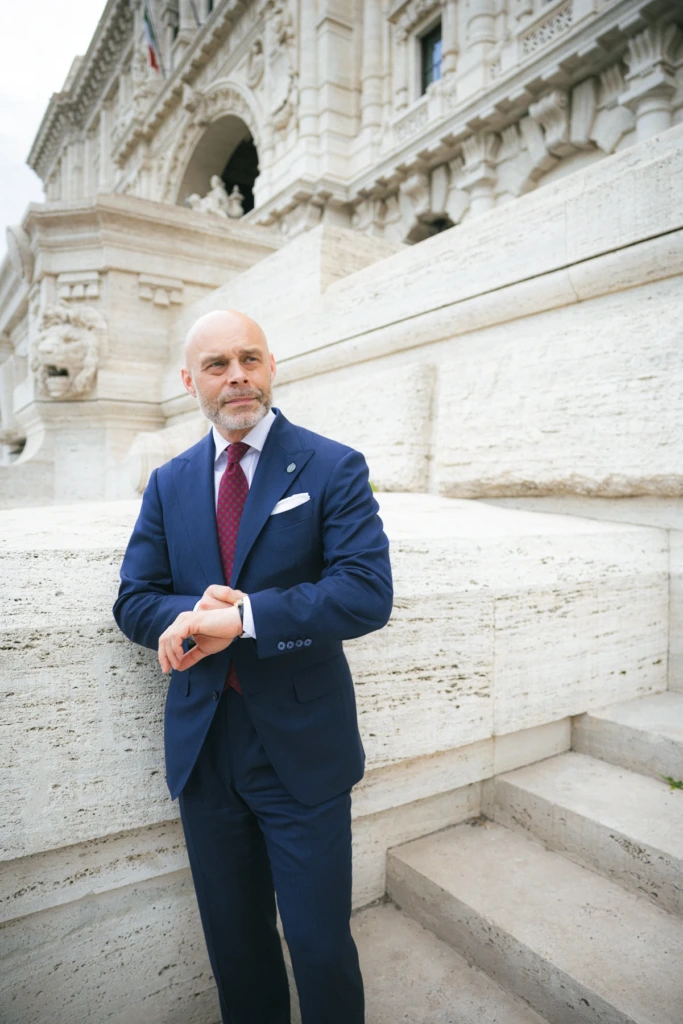 Mężczyzna w granatowym garniturze biznesowym, zapinający marynarkę, w białej koszuli i bordowym malinowym krawacie przed Sądem Najwyższym w Rzymie.
